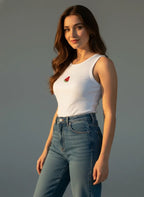 Woman wearing a white tank top with a red flower design and blue jeans against a gray background