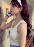 Woman in a white tank top with a colorful design, wearing a headband and Nike glove, standing in a locker room.