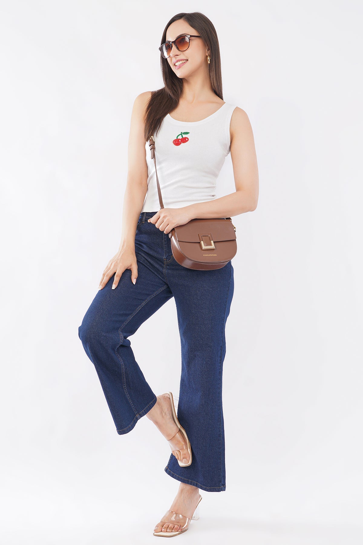 Woman wearing a white tank top with a cherry design and blue jeans, holding a brown handbag on a white background.